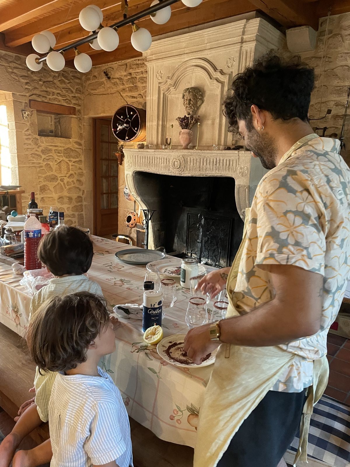 A guest rimming glasses with sumac salt in a stone farmhouse kitchen, kids watching